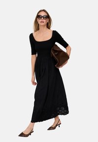 Schwarzes Maxikleid mit figurbetontem Oberteil, kurzen Ärmeln und besticktem Saum. Das Model hält eine braune Wildledertasche und trägt Schuhe mit Leopardenmuster.