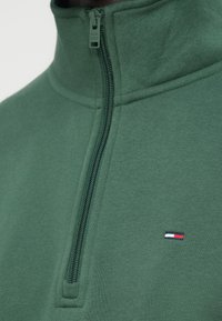 Πράσινη μπλούζα με ψηλό γιακά, που διαθέτει φερμουάρ μπροστά και μικρό λογότυπο σε κόκκινο, άσπρο και μπλε στη αριστερή πλευρά. Μαλακή υφή.