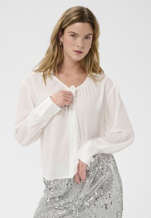 Femme aux cheveux blonds portant un chemisier blanc à boutons avec un lien au col, associé à une jupe argentée à paillettes, sur un fond uni.