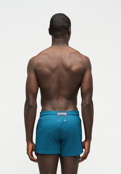 Άνδρας μοντέλο εικονίζεται από πίσω φορώντας μπλε πράσινα μαγιό με το λογότυπο Vilebrequin στη ζώνη, μπροστά σε απλό ανοιχτό φόντο.