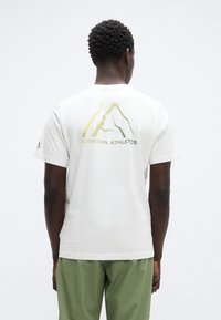 Λευκό βαμβακερό μπλουζάκι με χρυστό γραφικό βουνό και κείμενο "MOUNTAIN ATHLETICS" στην πλάτη. Κοντά μανίκια, άνετη γραμμή.