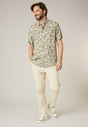 Homme souriant, portant une chemise à manches courtes verte à motif de palmiers, un pantalon beige et des baskets beige et blanches, debout devant un fond uni.