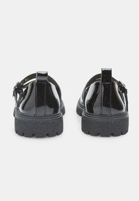 Zapatos de charol negro con una suela gruesa y texturizada, que presentan un diseño con el talón abierto y adornos decorativos en los lados.