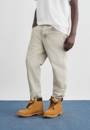 Persona con vaqueros beige y botas de trabajo marrón claro sobre una alfombra azul, con fondo gris claro.