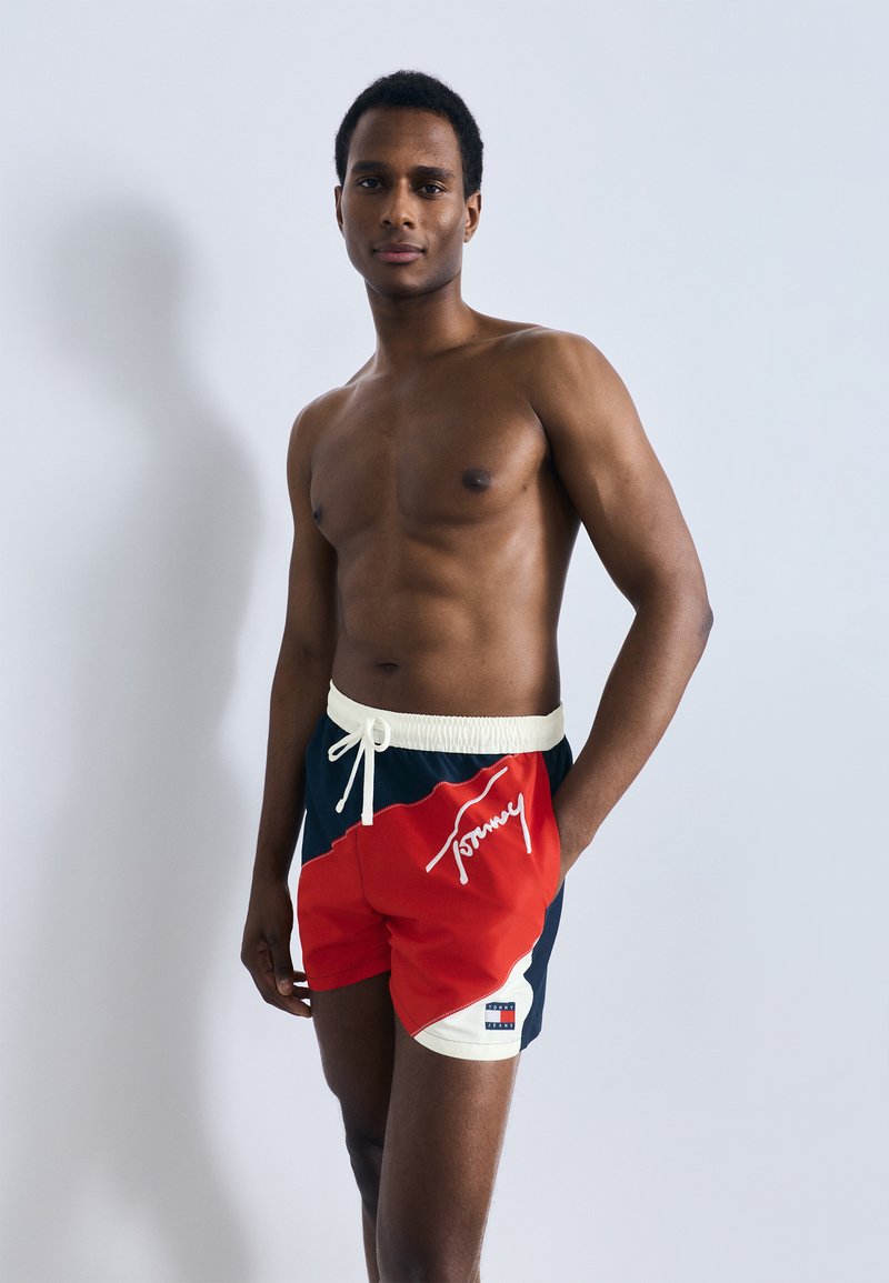 Κόκκινα και ναυτικά μαγιό σορτς με λευκή ζώνη μέσης, διακοσμημένα με κεντημένο λογότυπο και αντίθετες λεπτομέρειες, κατασκευασμένα από ελαφρύ ύφασμα που στεγνώνει γρήγορα.