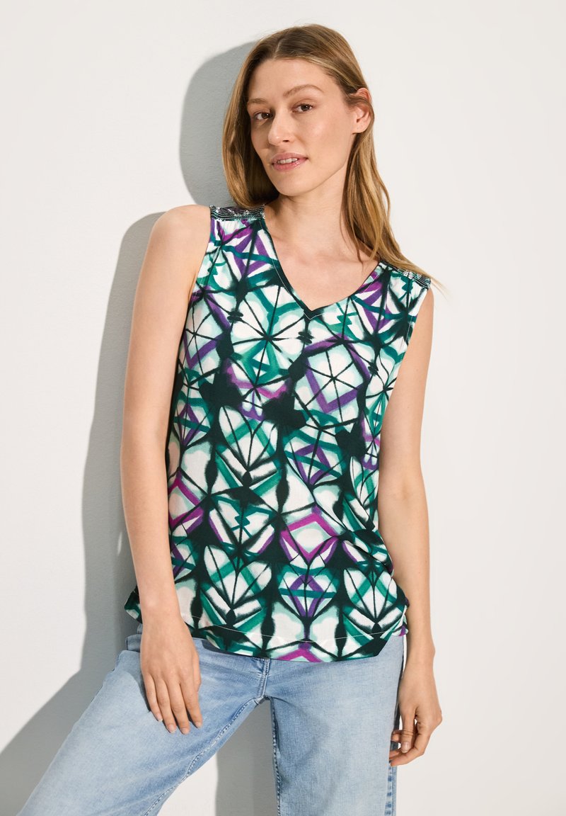 Top senza maniche con scollo a V, caratterizzato da un motivo geometrico in teal, bianco e viola su un tessuto leggero. Abbinato a jeans azzurri.