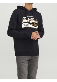 Felpa nera con grafica frontale in testo bianco e oro, dotata di cappuccio con cordini, polsini e orlo a coste. Indossata con jeans grigi.