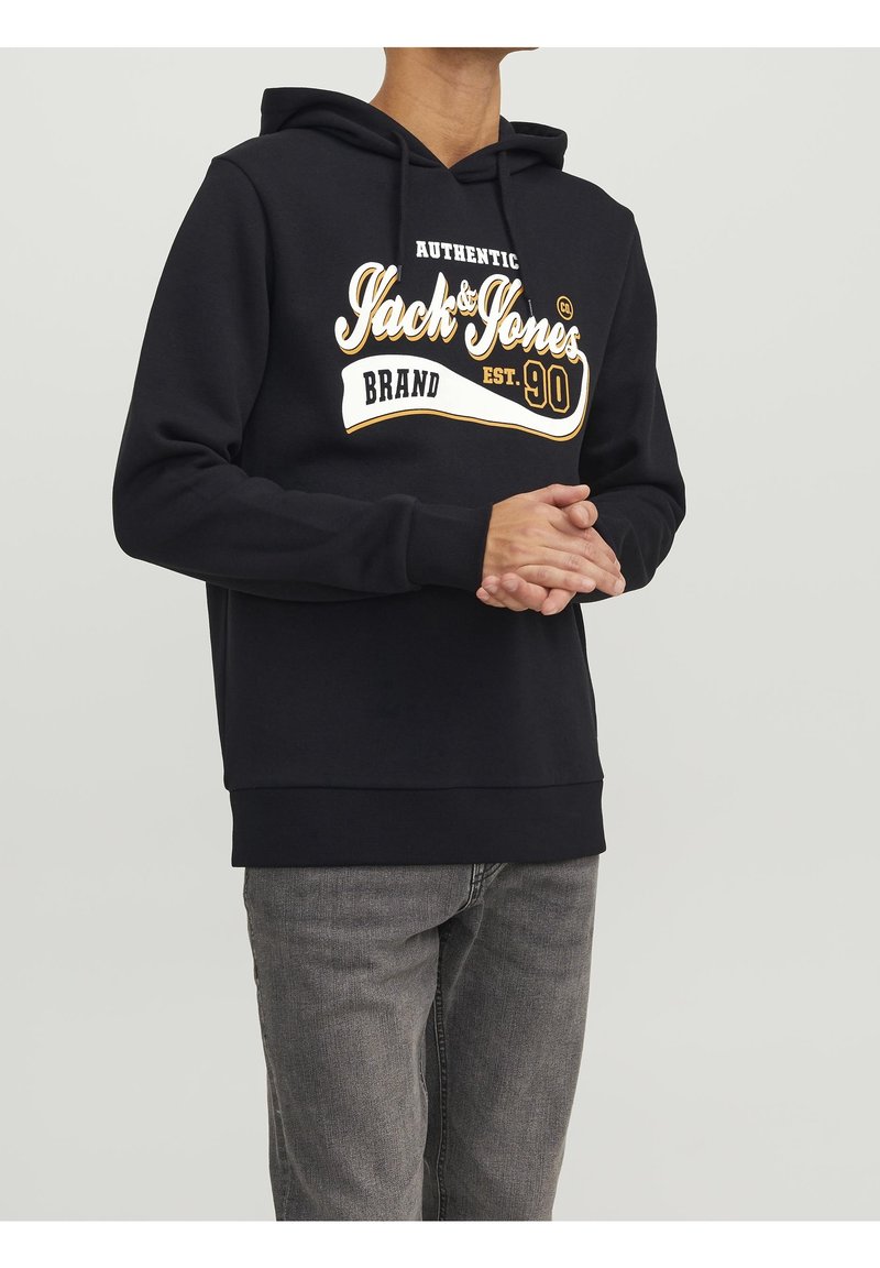 Felpa nera con grafica frontale in testo bianco e oro, dotata di cappuccio con cordini, polsini e orlo a coste. Indossata con jeans grigi.