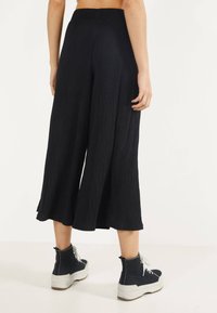 Black, wide-leg pleated trousers with a high waistband, featuring a textured fabric and a flowing silhouette. Worn with dark high-top shoes.