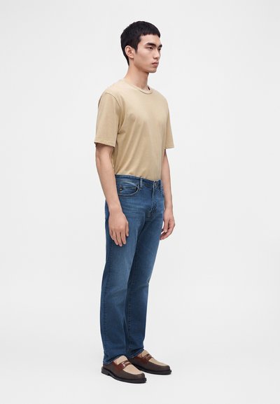 Homme portant un t-shirt beige, un jean bleu et des chaussures tissées marron, debout devant un fond uni gris clair, faisant face à droite.