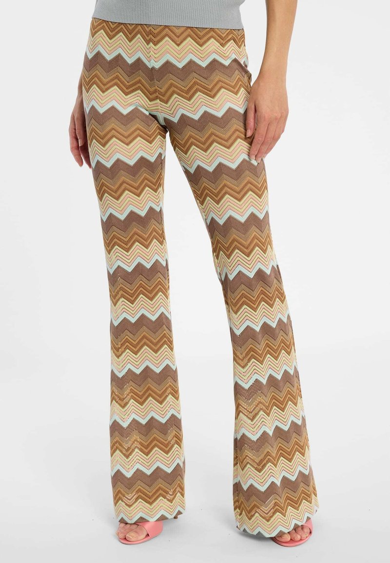 Persoon draagt bruine, beige en lichtblauwe flared broek met zigzagpatroon en roze open-toe sandalen, staat met één hand op de dij rustend.