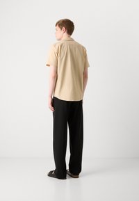 Kurzarm-Hemden mit Kragen in hellem Beige, kombiniert mit lockeren schwarzen Hosen, die einen entspannten Schnitt und ein minimalistisches Design zeigen.