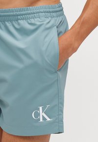 Lyseblå badebukser med elastisk linning, sidelommer og hvitt "CK"-logo på venstre ben. Laget av glatt, vannavstøtende materiale.