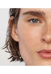 Gros plan sur un visage avec des cheveux courts et mouillés. Sourcils épais et noirs bien marqués, yeux clairs et bleus, peau lisse avec une texture naturelle et de légères taches de rousseur.