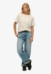 Camiseta beige de manga corta con logo en relieve; jeans azul claro, holgados y de pierna recta; zapatos negros con suelas texturizadas; puños enrollados.