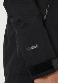 Gros plan de la manche noire de la veste O'Neill Hyperdry avec une sangle réglable en velcro et un détail du logo.
