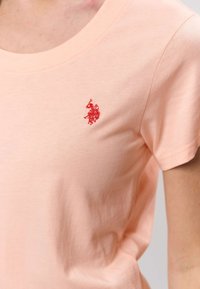 Persikanvärinen puuvillainen t-paita, jossa pyöreä pääntie ja lyhyet hihat, vasemmassa rinnassa pieni punainen kirjailtu logo.