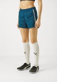 Pantaloni sportivi teal con cintura elastica, zip laterale bianca, abbinati a calze bianche fino al ginocchio e scarpini da calcio neri.