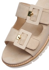 Beige ruskindsandal med to justerbare brede remme med store rektangulære cremefarvede spænder og hvid tekstureret sål.