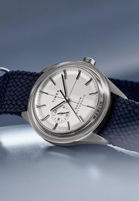 Stříbrné hodinky s kulatým pouzdrem z nerezové oceli, bílým ciferníkem a tmavě modrým tkaničkovým páskem. Mají zobrazení data a jednoduché značky hodin.