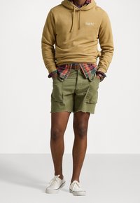 Felpa color marrone con tasca frontale e logo, indossata sopra una camicia a quadri. Pantaloni cargo verde oliva con orlo sfilacciato. Sneakers bianche completano il look.