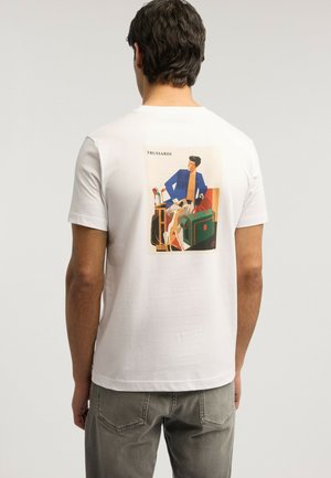 Uomo con maglietta bianca con grafica colorata sul retro raffigurante una persona, un cane, un bagaglio e mazze da golf, abbinata a pantaloni grigi.