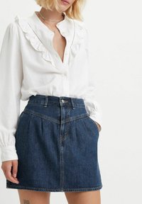 Blouse blanche avec col volanté associée à une jupe en denim foncé dotée d'une taille haute, de plis et de détails de couture visibles.