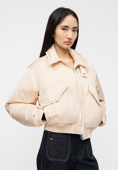 Broskyňovo sfarbená prešívaná bomber bunda s veľkým golierom, dvoma prednými vreckami a rebrovaným lemom. Nosená s tmavými džínsami s viditeľným šitím.