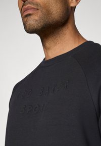 Tamsiai mėlynas sportinis megztinis su apvaliu kaklu, papuoštas tekstūruota, įspausta logotipo detalizacija ant krūtinės ir raglaniniu peties siūlėmis.