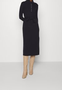 Lång marinblå klänning i mjukt tyg, med krage, knappdetaljer och ett bälte runt midjan. Kombinerad med knähöga, ljusbeiga stövlar.