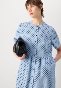 Abito azzurro con pois, maniche corte, chiusura a bottoni e gonna svasata. Clutch nera in evidenza. Texture liscia.