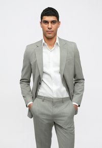 Ανοιχτό γκρι κοστούμι με διακριτές ρίγες, υφασμάτινη υφή, κλείσιμο με ένα κουμπί, σε συνδυασμό με λευκό πουκάμισο, χέρια στις τσέπες, ουδέτερο φόντο.