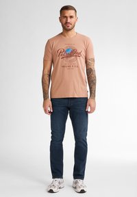 Pánské světle růžové bavlněné tričko s grafickým potiskem v tmavě hnědé barvě, spárované s tmavě modrými slim-fit džíny a bílými teniskami.