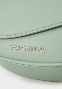 Borsa in pelle verde dalla forma arrotondata, con un bordo cucito e un logo "Valentino" in oro nella parte inferiore. Texture liscia.