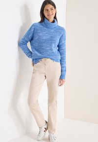 Femme en pull à col roulé bleu en tricot et pantalon beige, appuyée contre un mur blanc, souriante, portant des baskets blanches avec des accents rouges.