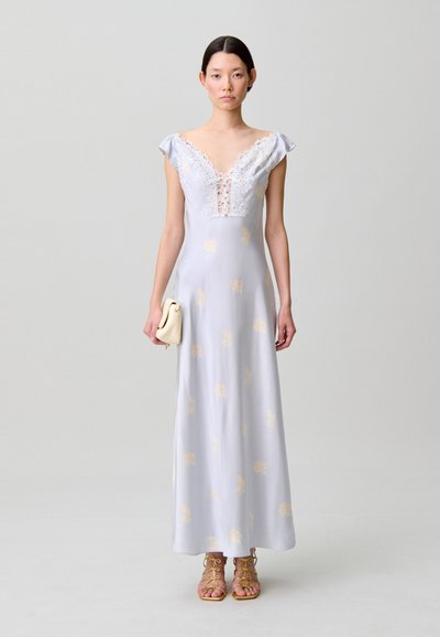 Donna che indossa un maxi abito floreale azzurro con scollo a V in pizzo, che tiene una pochette beige e indossa sandali gialli con cinturini su uno sfondo neutro.