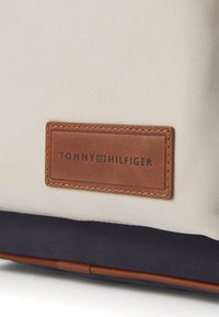 Borsa in tessuto beige con fondo blu navy; presenta un'etichetta in pelle marrone impressa con "TOMMY HILFIGER" e una sottile texture a righe diagonali.