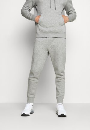 Persoon draagt een grijze trainingsbroek en een bijpassende hoodie met de handen in de voorzak, staand met witte sportschoenen aan.