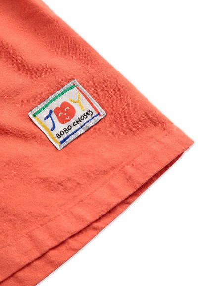 Korallfarbenes Kurzarmhemd mit einem Stoffetikett, das "JOY" und ein lächelndes Apfeldesign zeigt, umrandet von bunten Rechtecken.