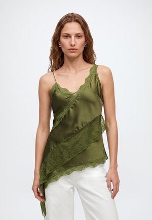Femme aux cheveux ondulés portant un caraco en satin vert bordé de dentelle et un pantalon blanc, debout devant un fond clair uni.