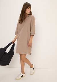Femme en robe beige mi-longue et baskets blanches tenant un grand sac fourre-tout noir, regardant sur le côté devant un fond uni.