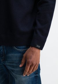 Marineblauwe gebreide trui met ribgebreide manchetten, voorzien van een logotag. Gecombineerd met blauwe jeans met een casual fit en subtiele slijtplekken.