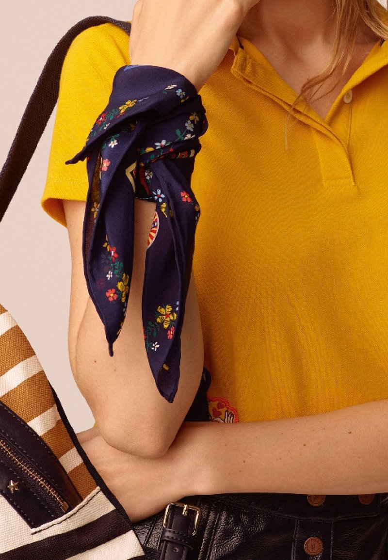 Écharpe en soie marine avec des motifs floraux nouée autour d'un bras, portée avec un polo jaune. Comprend également un sac à main rayé en arrière-plan.