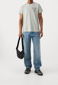 Vit t-shirt med kort ärm och grafiskt tryck på framsidan, lösa blå denimjeans och en svart axelväska. Grå sportskor med svarta detaljer.