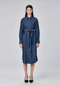 Femme se tenant dans une robe en denim bleu marine à boutons, longueur genou, à manches longues, avec une ceinture nouée à la taille et des talons noirs pointus.