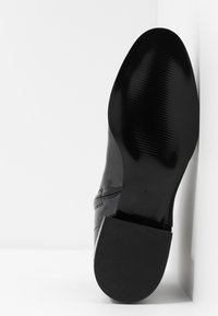 Zapato de charol negro con punta redonda, suela texturizada y acabado brillante. Presenta detalles de costura sutiles a lo largo de la parte superior.