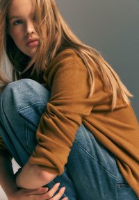Brązowy sweter o teksturowanej tkaninie i podwiniętych rękawach; niebieskie dżinsy w luźnym fasonie. Model znajduje się w pozycji kucającej.