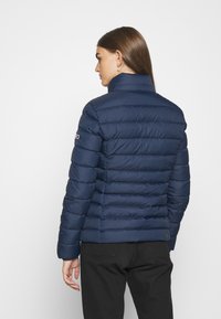 Chaqueta acolchada azul marino con cuello alto, textura acolchada y un sutil detalle de logo en la manga. Presenta un diseño ajustado y mangas largas.