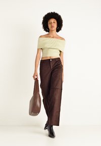 Světle zelený crop top s odhalenými rameny a ribbed texturou, hnědé široké kalhoty s předními kapsami, doplněné hnědou kabelkou přes rameno a černými botami na podpatku.