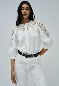 Blusa blanca con detalles de encaje en los hombros, botonadura frontal y mangas abullonadas de 3/4. Combinada con pantalones blancos y un cinturón negro. Textura suave.